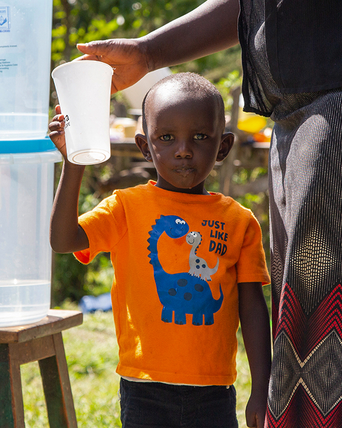 A young child wearing an orange shirt with dinosaurs holds a clear plastic cup up as an adult pours water from a large dispenser; outdoors on a sunny day.