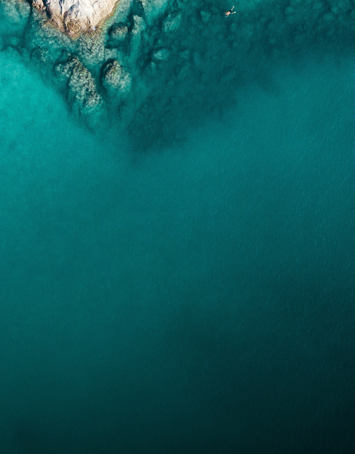 aerial view of a rocky coastline meeting clear turquoise water, with lighter shallow areas near rocks at the upper left and a lone swimmer visible in the sea.