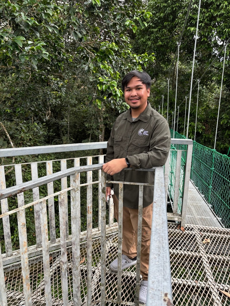 A person standing on a metal walkway wearing a green button-up shirt and tan pants, leaning on a railing with a lush green forest in the background.