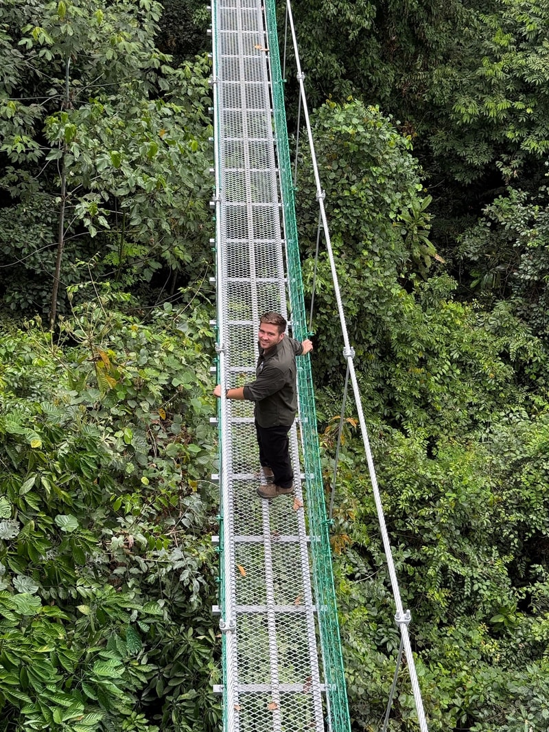 A person stands on a narrow metal suspension bridge high above dense green foliage, holding onto the railing and smiling as they cross the bridge.