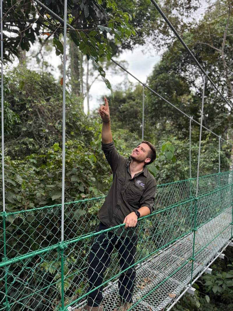 A man stands on a metal mesh suspension bridge in a dense green forest, pointing upward with one hand while leaning on a green chain-link railing.