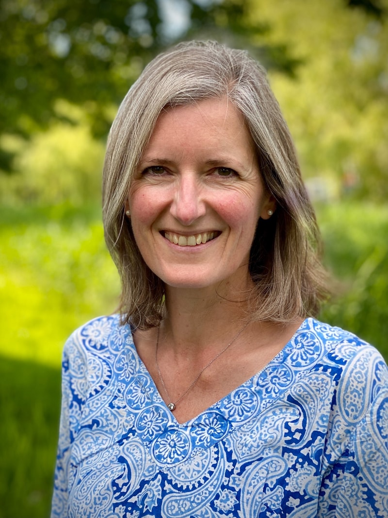 Smiling woman with shoulder-length gray hair wearing a blue and white patterned top, standing outdoors in a green, sunlit park background.