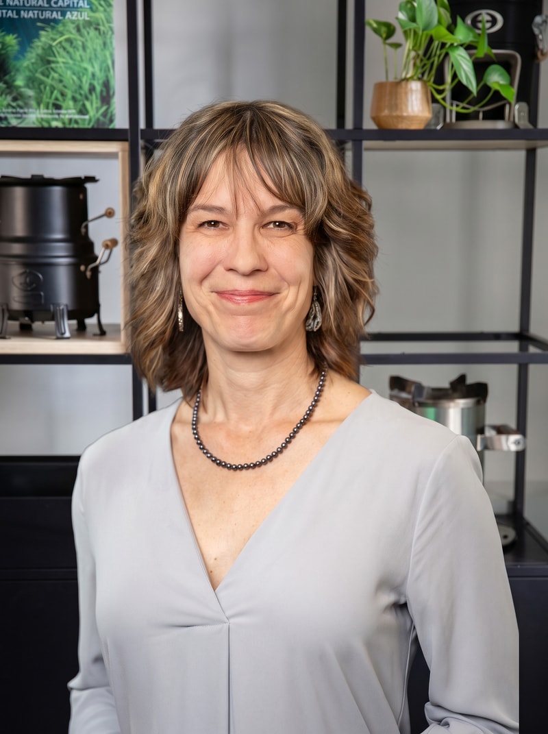 Smiling woman with shoulder-length light brown hair and bangs, wearing a gray blouse, a beaded black necklace, and dangling earrings, standing in front of a shelving unit with plants and equipment.