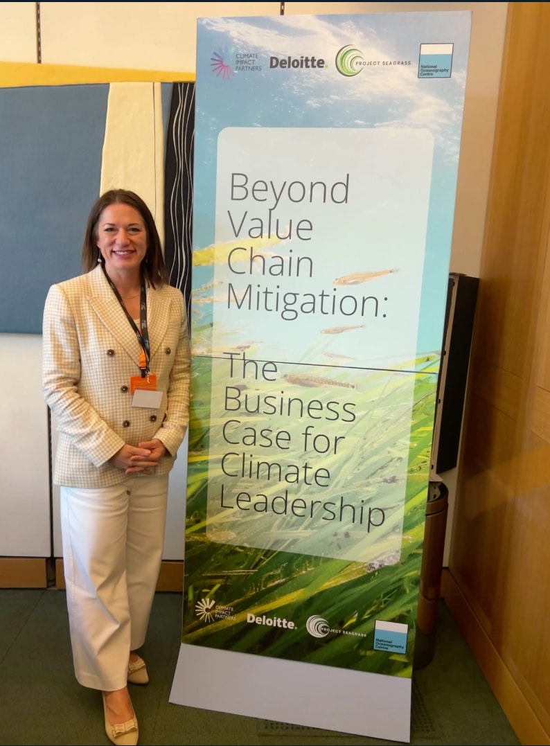 A smiling woman in a light blazer and white pants stands beside a tall conference banner that reads “Beyond Value Chain Mitigation: The Business Case for Climate Leadership” with logos at the top.