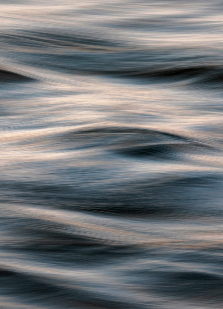 Waves or ripples on a body of water with soft, blurred motion creating horizontal bands of blue, gray, and beige hues, resembling abstract flowing patterns.