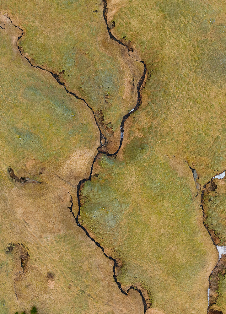 Aerial view of a dry, meandering crack through grassy terrain with brown and green patches, showing a winding fissure and small rocky edges.