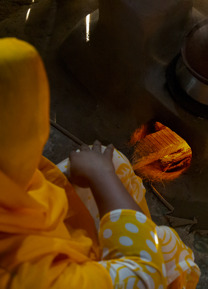 Person wearing bright yellow clothing sits near a lit clay oven, hands resting on a patterned fabric, warm orange glow from the fire illuminating the scene.