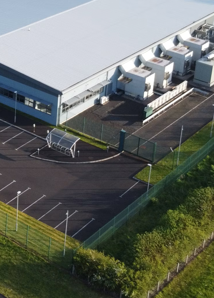Aerial view of a large industrial building with white exterior, multiple loading docks, a fenced parking lot, and surrounding green areas.