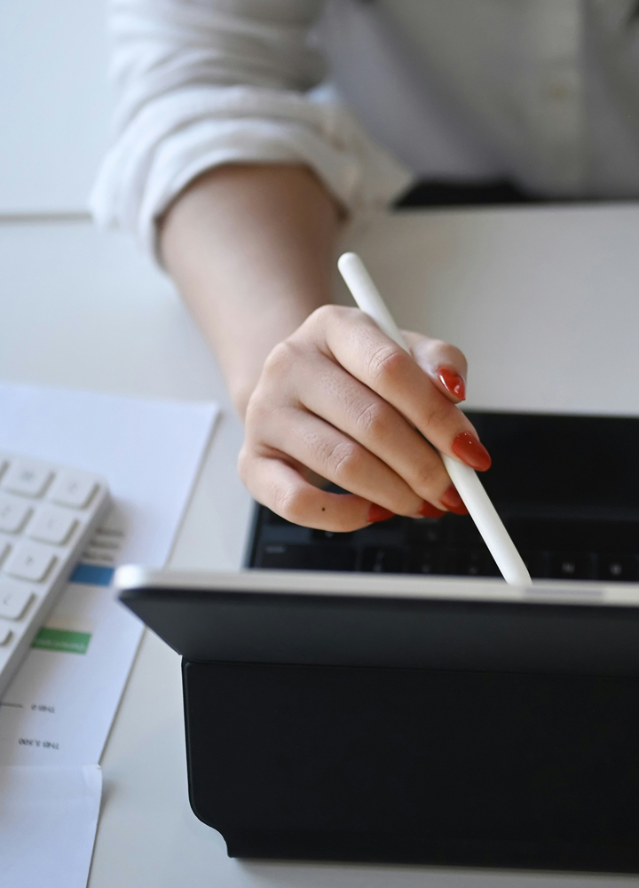 A person uses a white stylus on a tablet at a desk, with a keyboard and papers nearby; the focus is on the hand with orange nail polish.