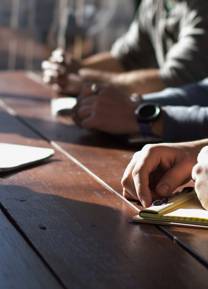 People sit around a wooden table outdoors, focused on writing or drawing with pencils, with hands and a yellow notebook in the foreground.