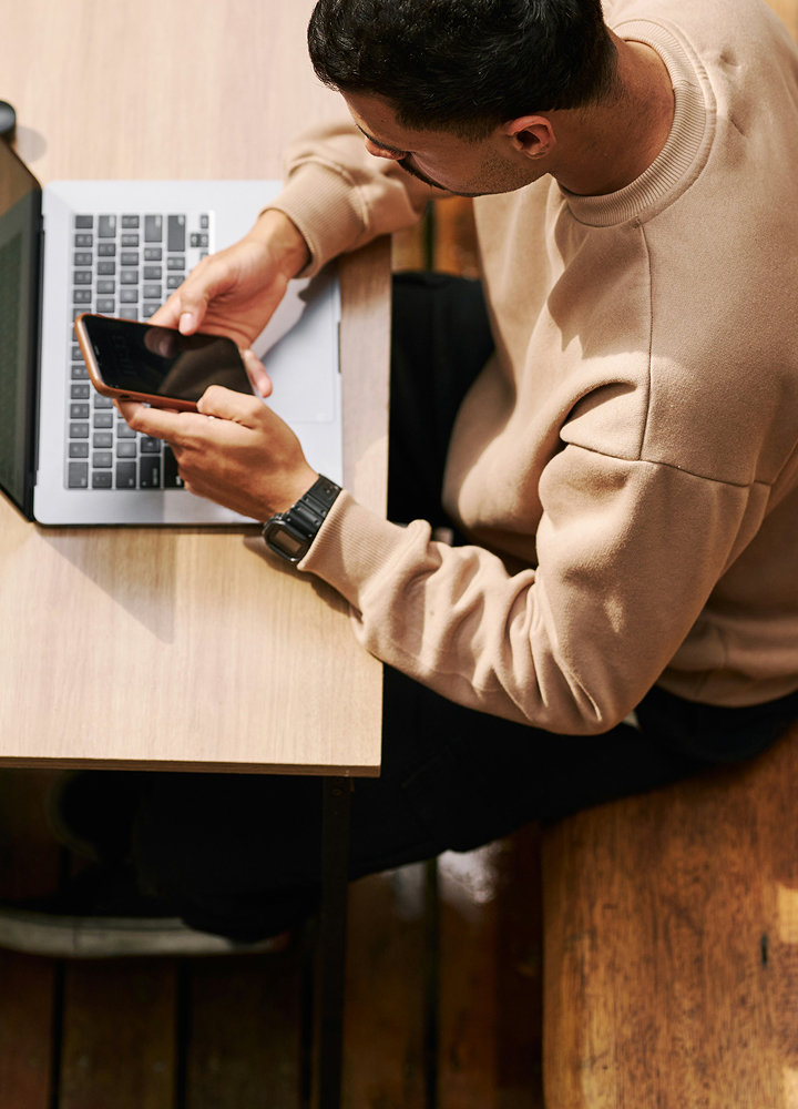 Person wearing beige sweatshirt sits at wooden desk, using a smartphone with one hand while the other rests near a laptop keyboard.