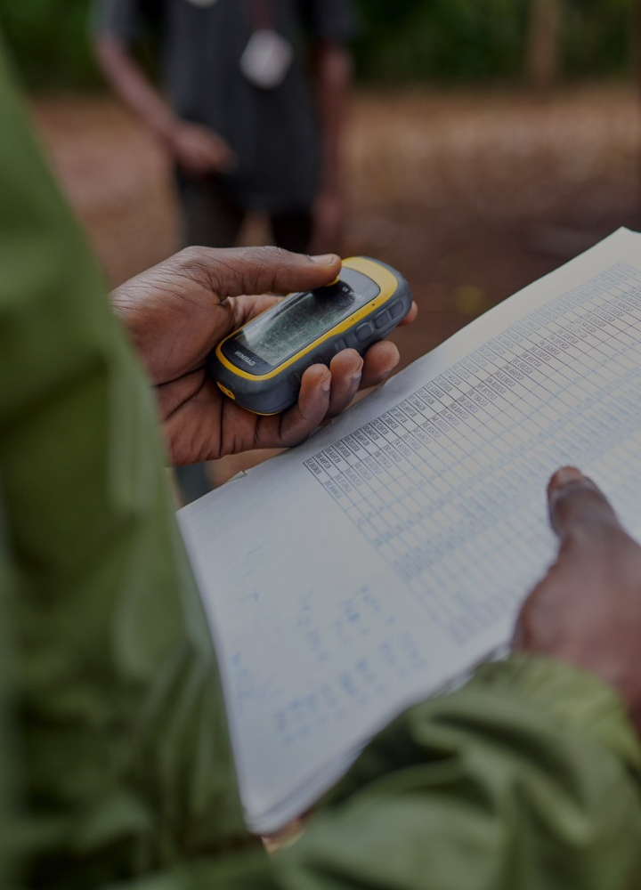 A close-up of a person holding a rugged handheld GPS device over a notebook with a data table, outdoors with a blurred background.