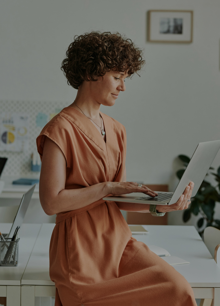 A person with short curly hair sits on a desk working on a laptop in a bright, modern office wearing a peach-colored dress.
