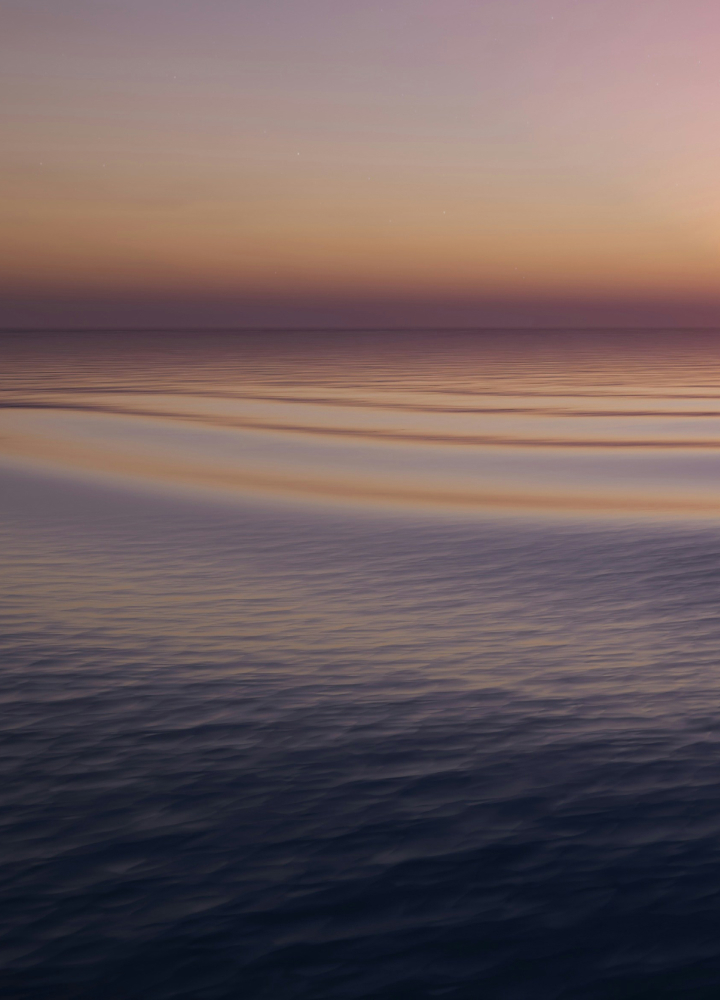A tranquil ocean scene at sunset with smooth water, soft purple and orange hues on the horizon, and gentle ripples reflecting the sky’s colors.