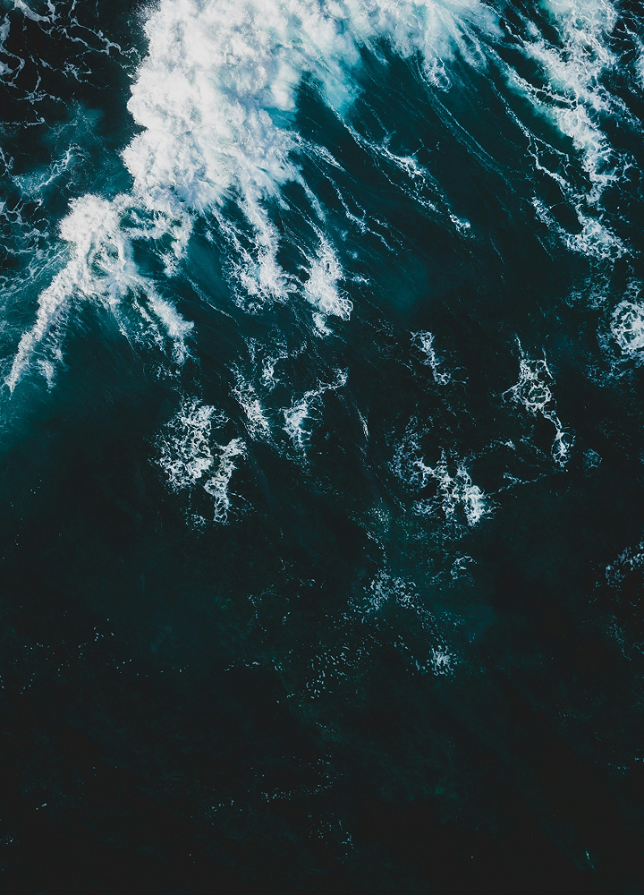Aerial view of dark teal ocean with white foamy waves crashing and swirling, creating turbulent, frothy patterns across the water.