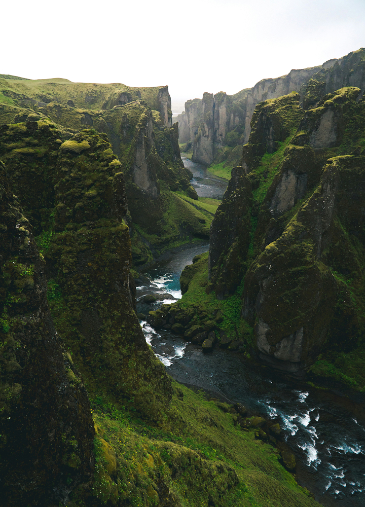 A dramatic canyon with steep mossy cliffs and a winding river cutting through, rocky ledges, and green vegetation along rugged gorge walls.
