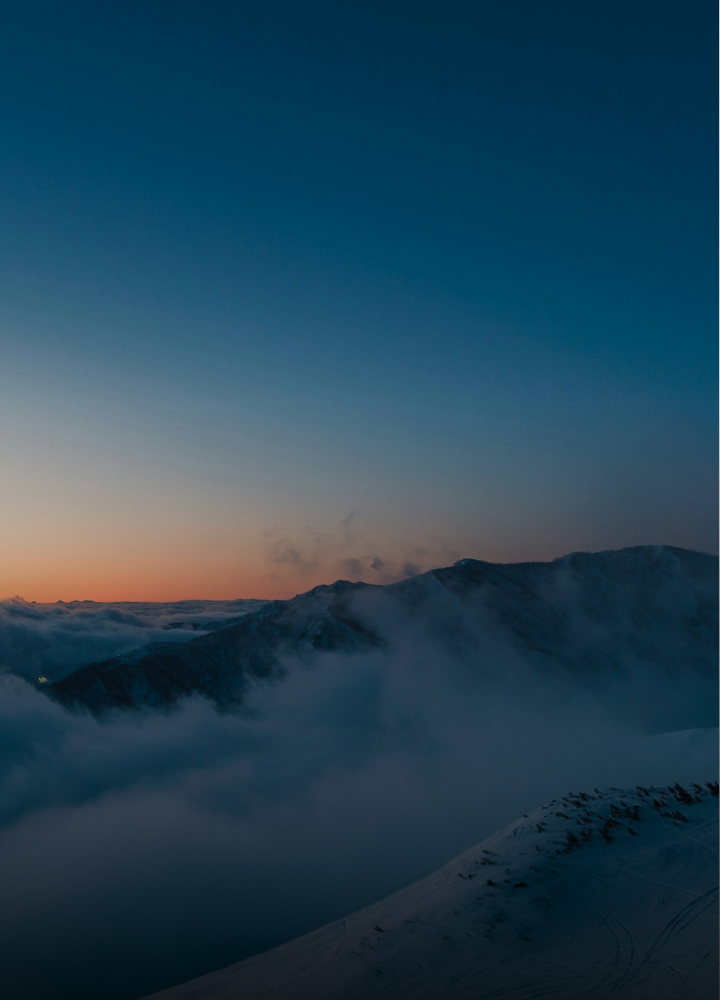 Daintily lit mountain ridge emerging through low-lying clouds at dusk, with a clear gradient sky from pale orange near the horizon to deep blue overhead.
