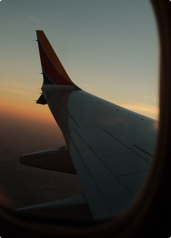 Sunset view of an airplane wing seen from a window, with a colorful winglet, soft orange and blue sky, and distant horizon below.
