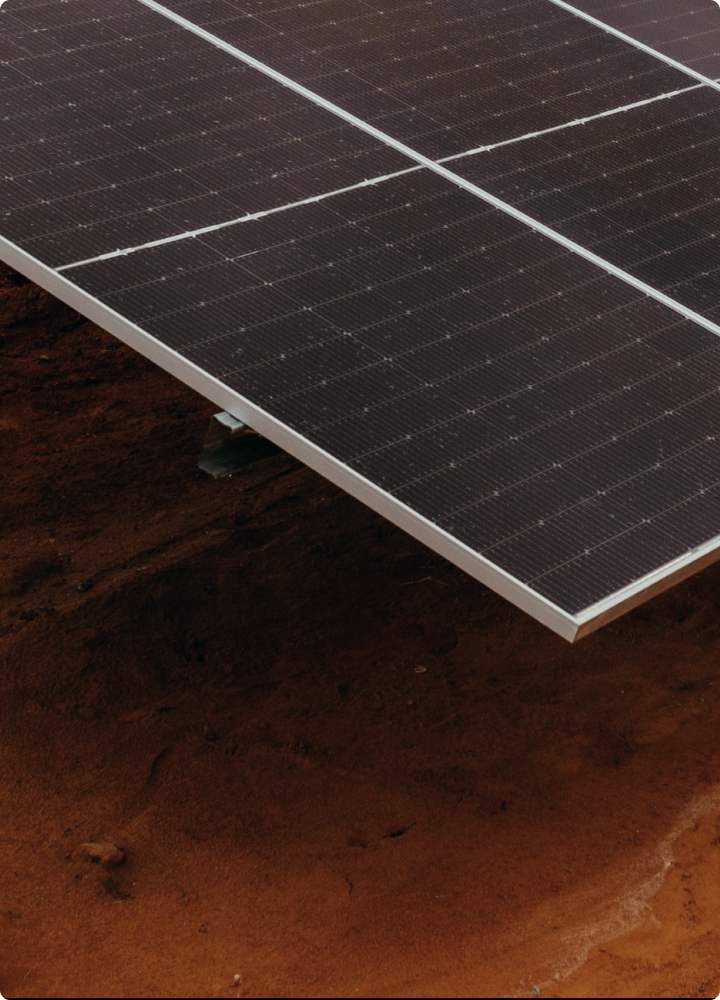 Solar panel tilted over reddish soil, close-up view showing dark photovoltaic cells with white grid lines, metal edge, and a supporting mount partly visible.