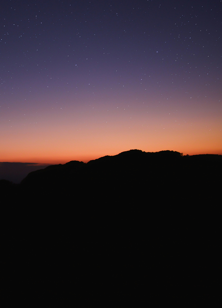 Stars fill a dark twilight sky above a silhouetted horizon and rugged hill at sunset, with a warm orange glow along the horizon fading to deep blue.