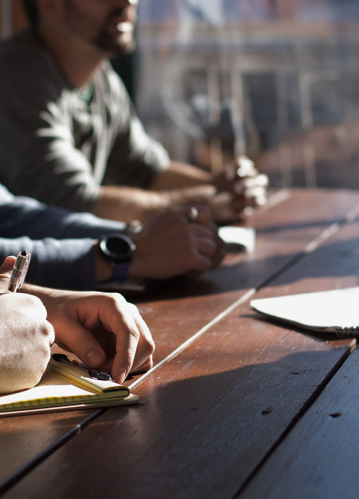 Several people sit at a wooden table, hands visible as one writes in a notebook with a pen, others hold objects and appear engaged in a discussion.