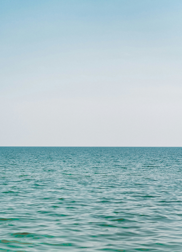Calm turquoise ocean under a clear pale blue sky, horizon line stretching across the middle with gentle waves lapping the surface.