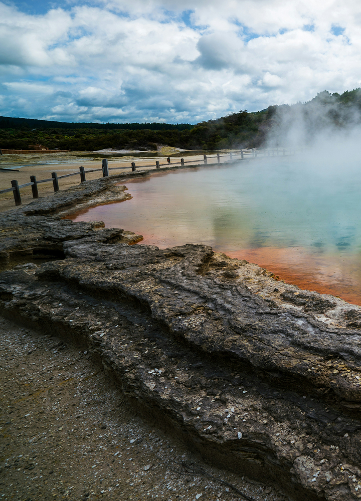Steam rises from a colorful hot spring with turquoise water and orange mineral rims; a weathered rock ledge and wooden fence line the edge under a cloudy sky.