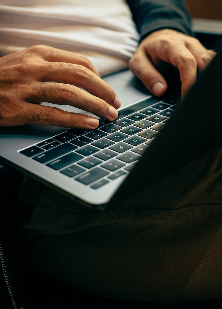 Hands typing on a laptop keyboard, close-up view of fingers over dark keys, metal trackpad area visible, person seated with casual clothing.