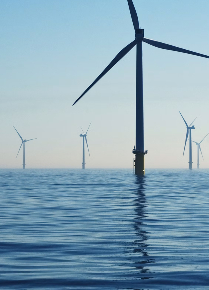 Offshore wind turbines rise from calm blue sea, their tall towers and rotating blades cutting through a misty horizon, with reflections shimmering on the water.