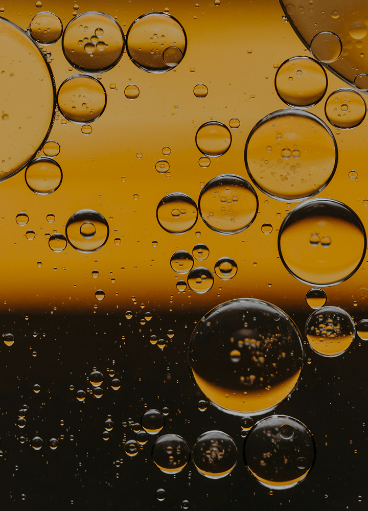 Oil bubbles suspended in a golden liquid, varying sizes with light reflecting off curved surfaces, against a warm yellow-to-dark gradient backdrop.