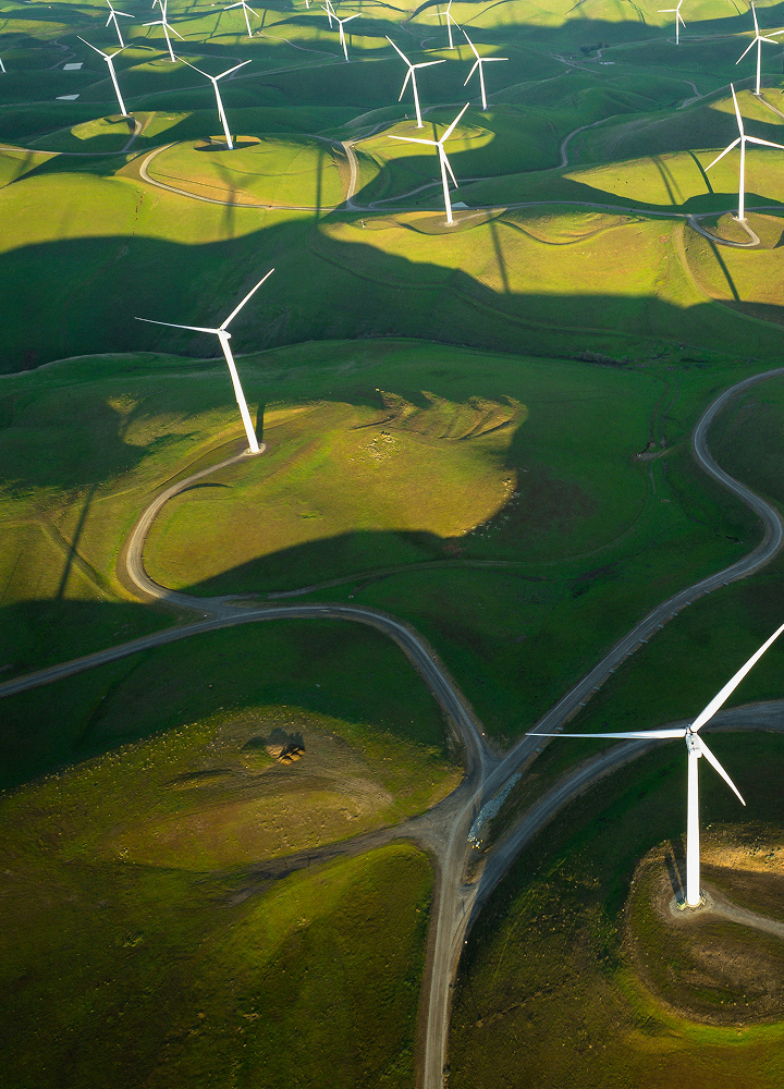 Aerial view of rolling green hills with numerous white wind turbines, winding roads connecting turbine bases and casting long shadows across the landscape.