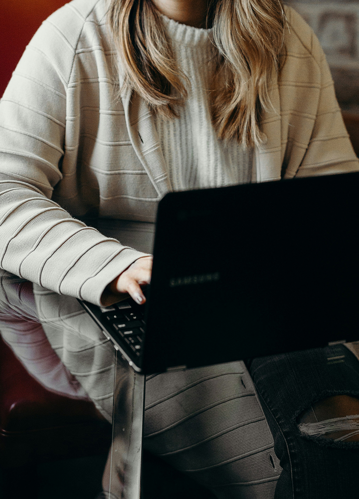 Person seated at a reflective glass table using a black laptop, wearing a light striped cardigan and a light knit sweater; blonde hair partially covering the screen.