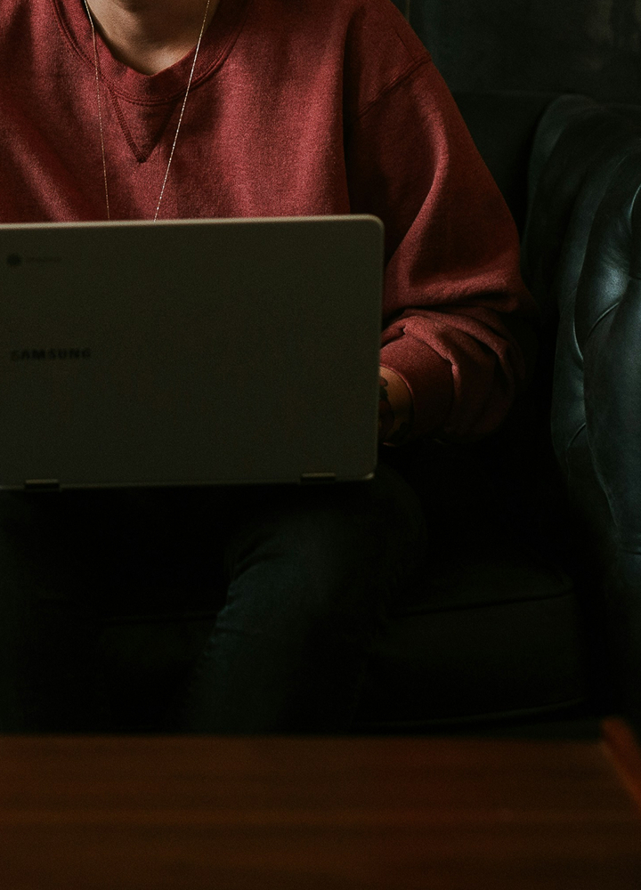 Person seated on a dark sofa using a Samsung laptop; wearing a red sweatshirt and a thin necklace, with the laptop resting on their lap in a dimly lit room.