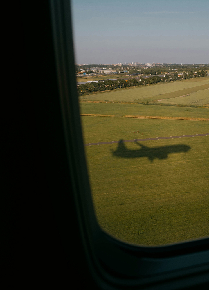 Shadow of a small airplane on a grassy airfield seen from an airplane window, with distant cityscape and blue sky in the background.