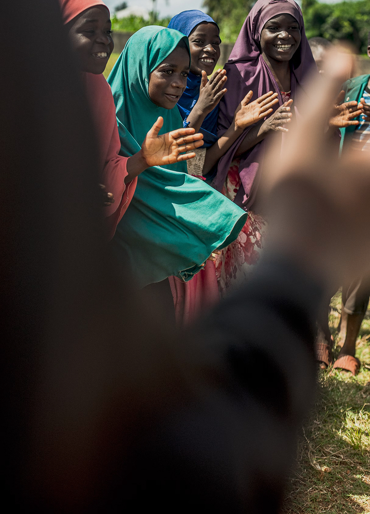 Group of smiling women in colorful hijabs outdoors, raising hands in a joyful gesture, with sunlit grass and blurred figures nearby.