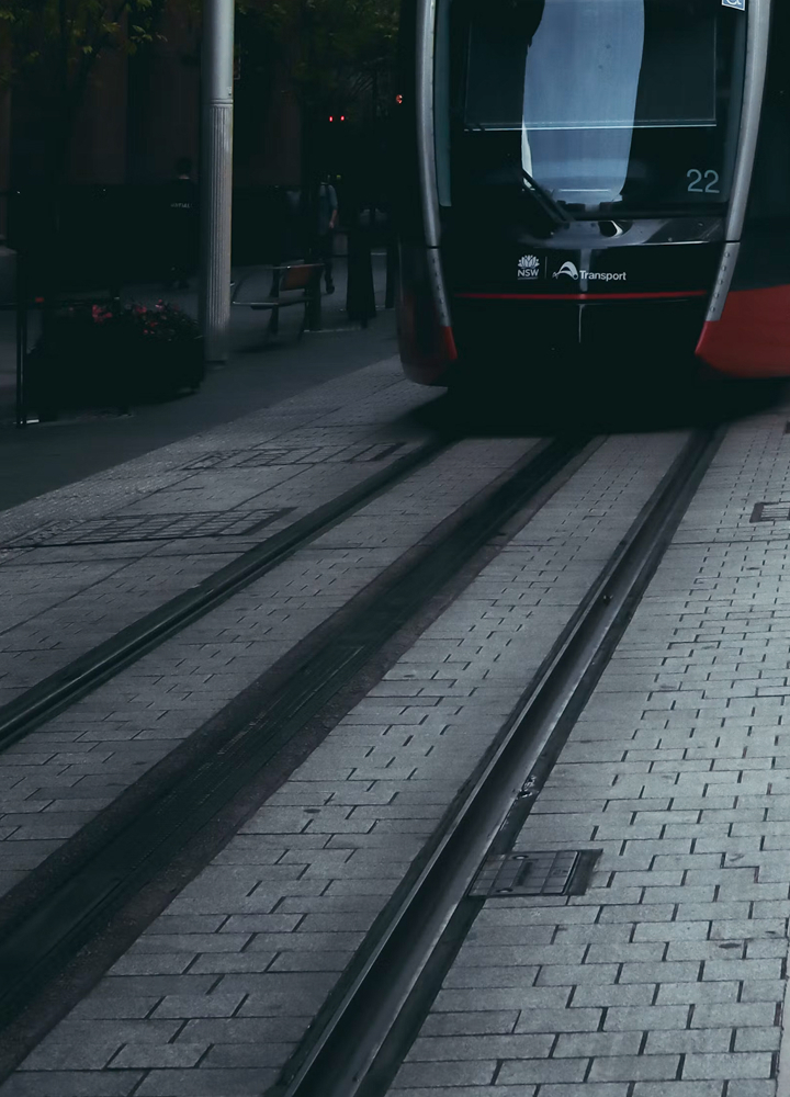 Light rail tram approaching on tracks in an urban plaza with paved sidewalk, rails, and a dark, shadowy background.