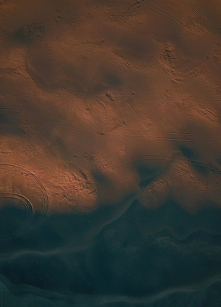 Desert terrain with reddish sand patterns and tire tracks, transitioning to dark blue shadows at the bottom, showing wind-eroded ridges and circular markings.