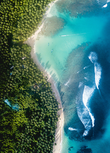 Bird’s-eye view of a tropical coastline where dense green forest meets a narrow white-sand beach, turquoise shallow waters transitioning to deep blue, with gentle waves and a few small boats.