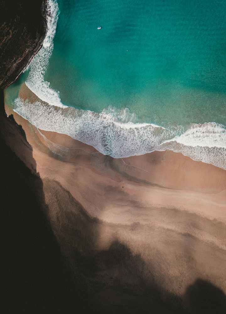 Aerial view of turquoise sea meeting a sandy beach with white surf, shadows from cliffs on the left, and a lone boat near the water's edge.