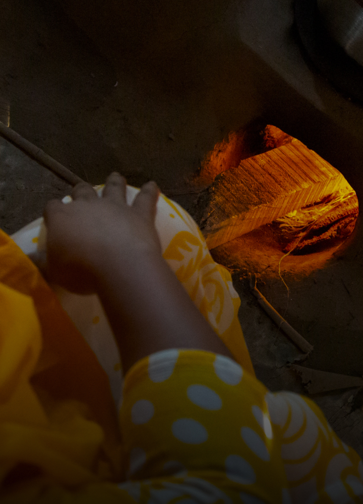 A hand in a yellow patterned garment reaches toward an opening in a wood-fired oven with orange glowing embers and a warm fiery interior.