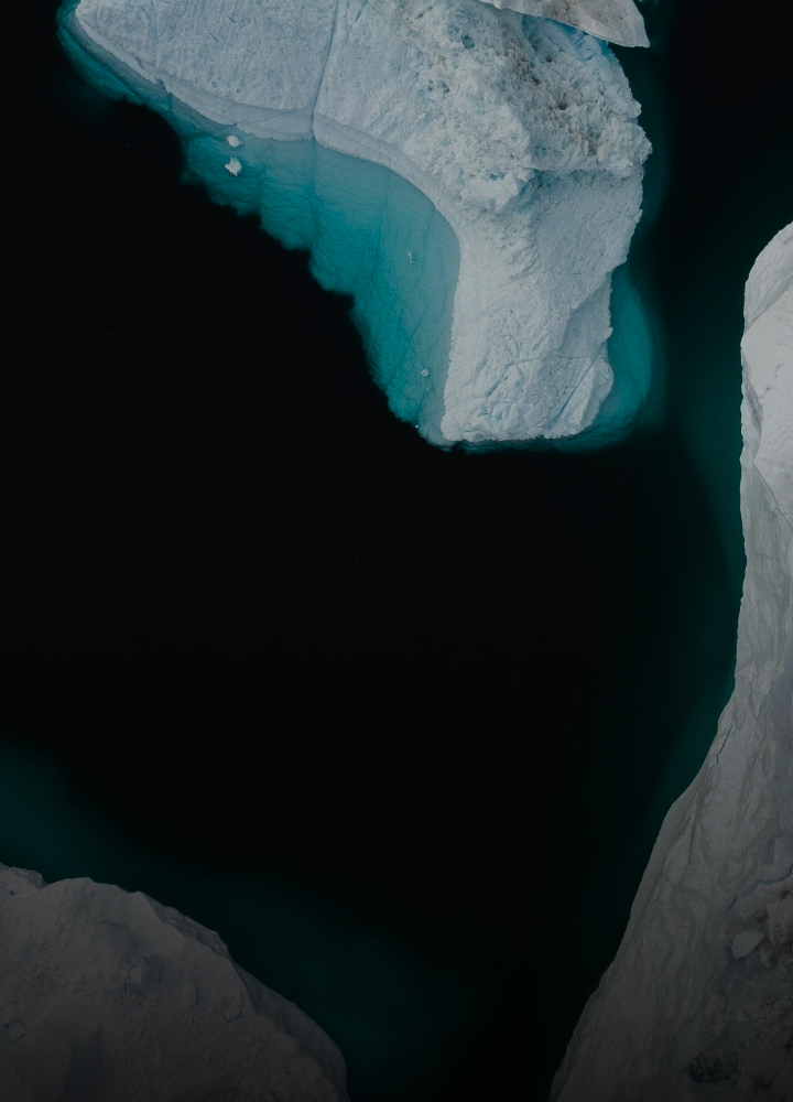 Aerial view of a dark, narrow waterway bordered by white ice cliffs with turquoise glints along the edges, forming a winding icy chasm.