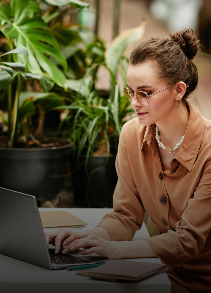 A woman with brown hair in a bun wearing glasses and a beige button-up shirt works on a laptop at an outdoor desk, surrounded by green plants.