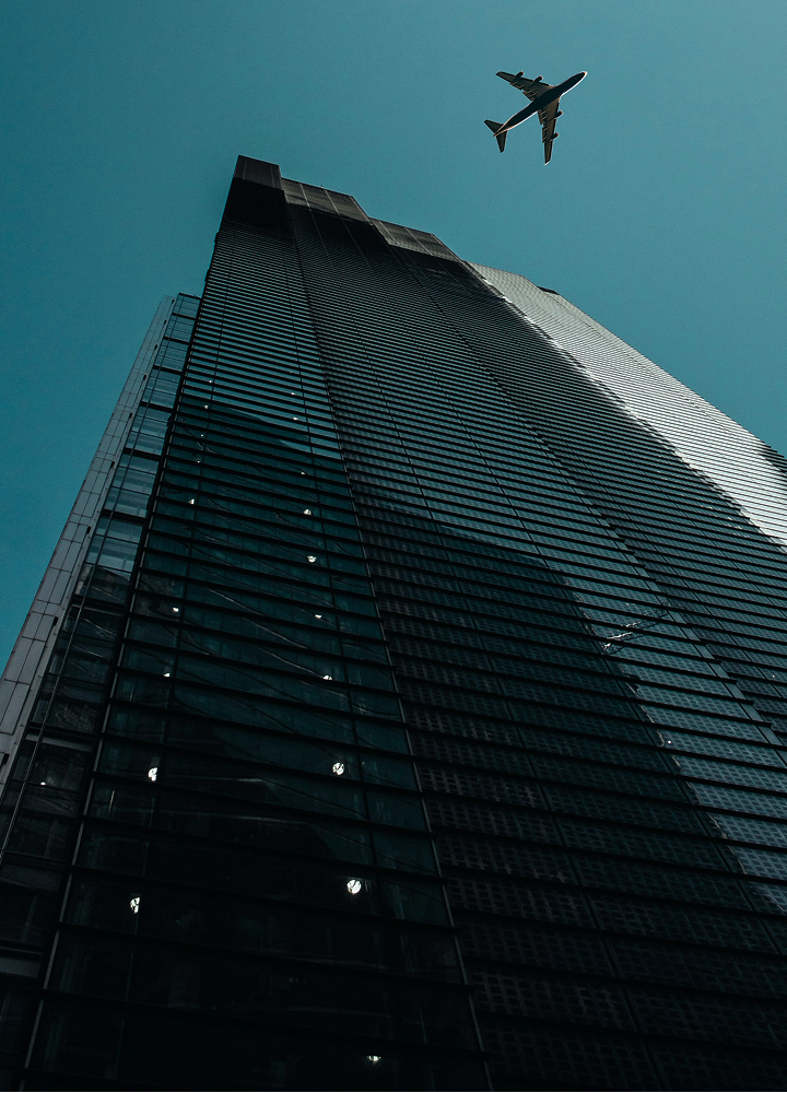 A tall glass skyscraper seen from below with reflections in its dark facade, and a passenger airplane flying overhead against a clear blue sky.