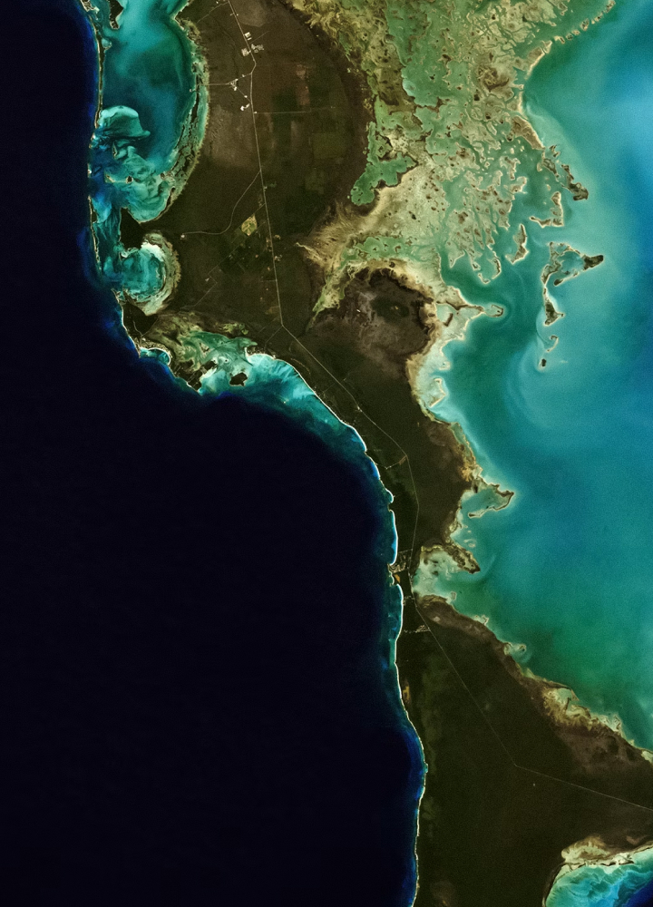 Coastal satellite view showing dark ocean to the left and turquoise reef waters along jagged shoreline with green land, roads, and small inland patches.