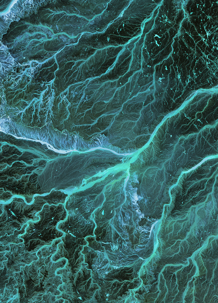 Intricate teal and turquoise river-like channels weave across a dark ocean surface, creating bright, veined patterns and feathered edges throughout the image.