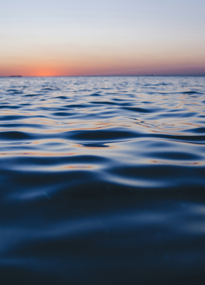 Serene seascape at sunset with gentle blue waves in the foreground and a warm orange-pink horizon fading into pale sky.