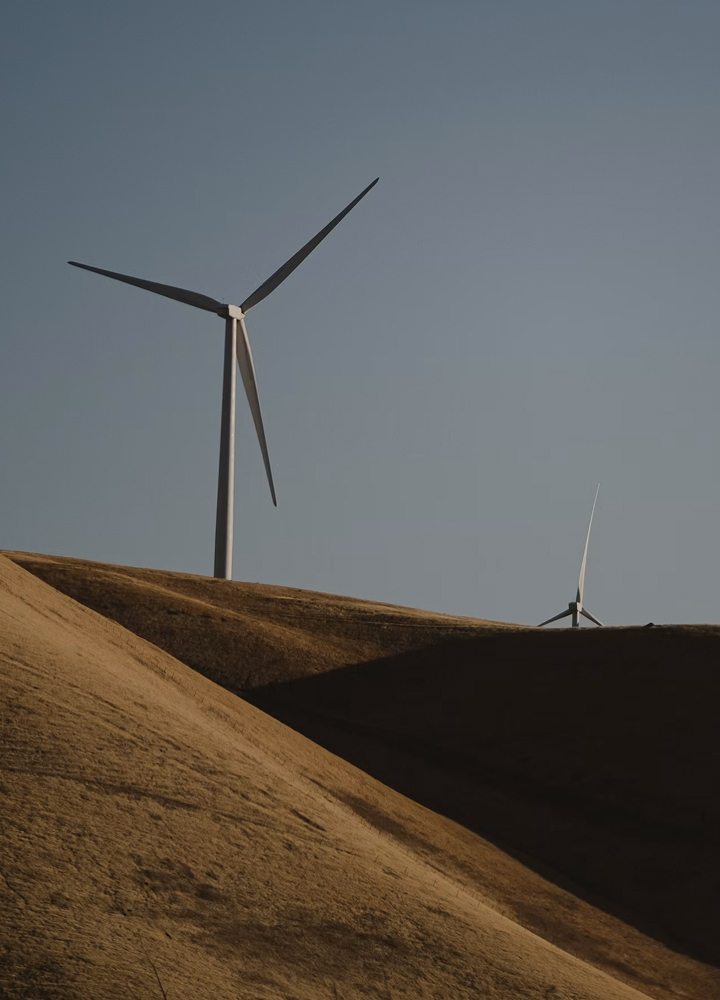 Two large wind turbines rise above rolling, sunlit brown hills under a clear blue sky, with a smaller turbine visible in the distance on the right.