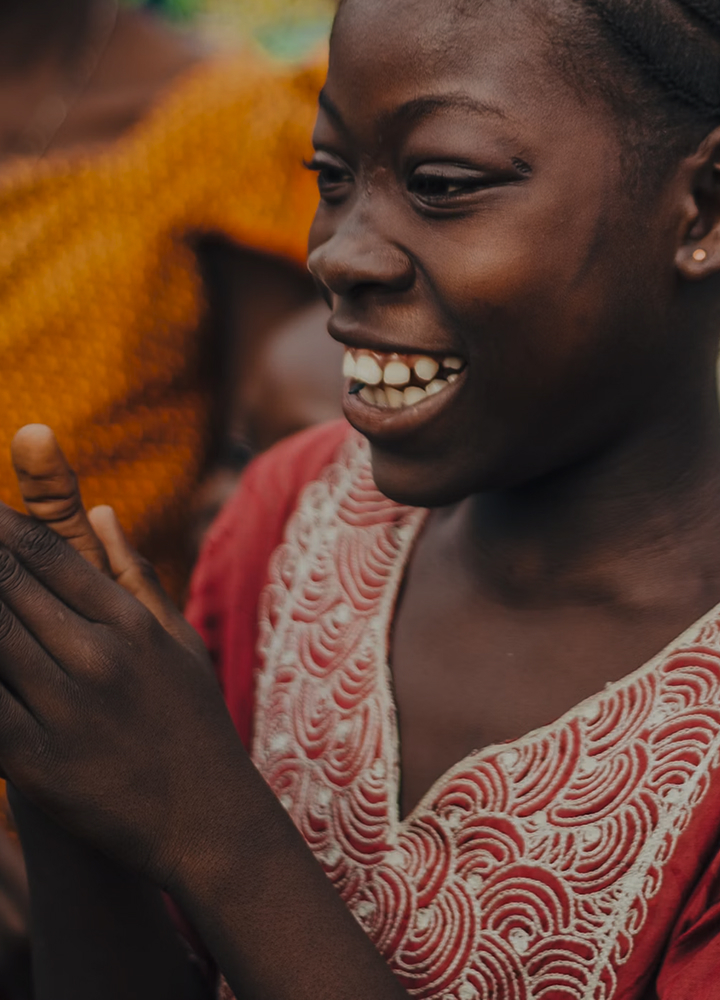 Close-up of a smiling person with dark skin, wearing a pink embroidered garment, showing teeth and a joyful expression, hands raised in the foreground.