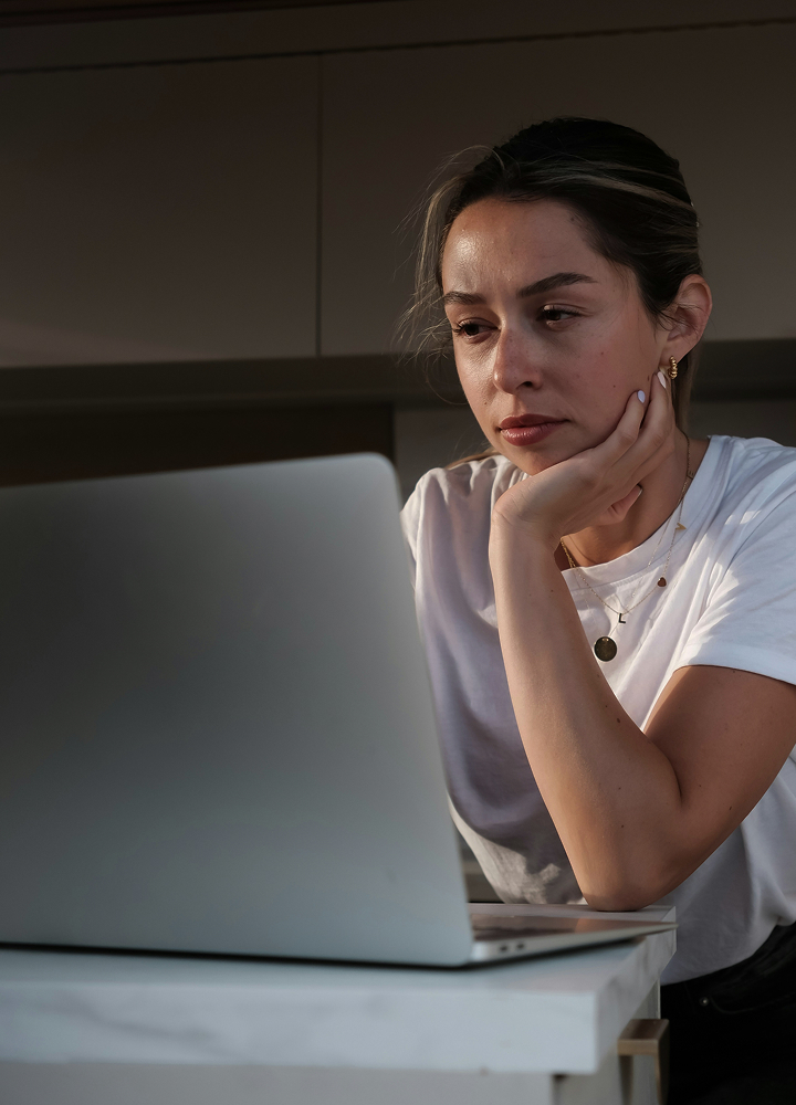 A woman in a white t-shirt sits at a desk, resting her chin on her hand while looking down at a laptop with a pensive expression.
