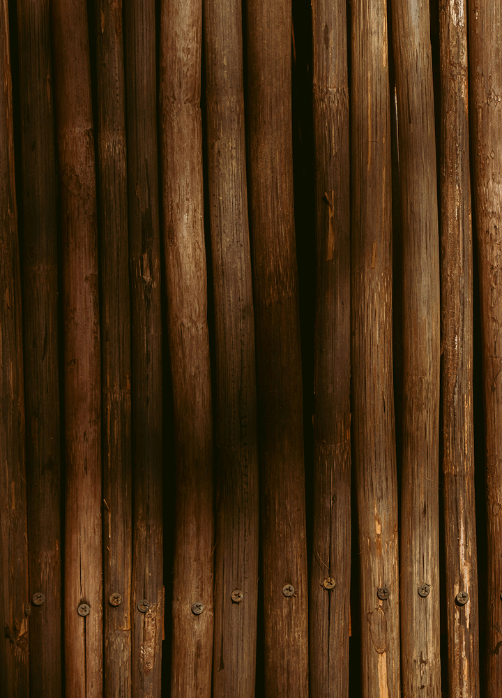 Stacks of weathered wooden logs or poles arranged vertically, dark brown tones with visible grain, knots, and rusty nails at the bottom.